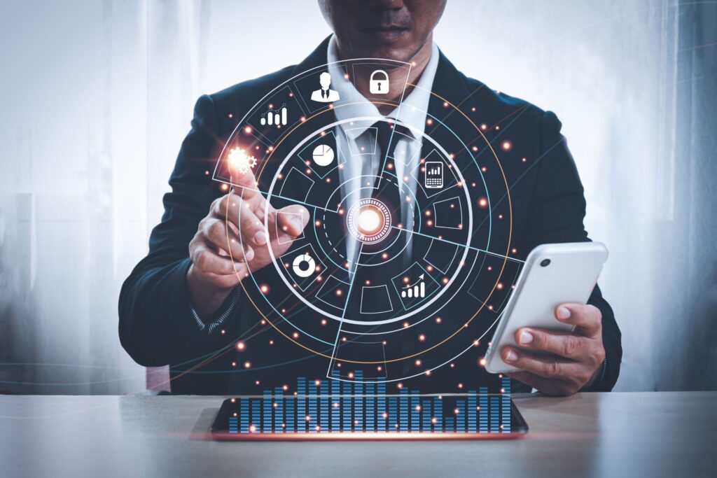 Asian man in a suit sitting at a desk with his left hand holding a mobile phone. His right hand pointing at a hologram showing business icons in hi-tech circle. Black background blur.